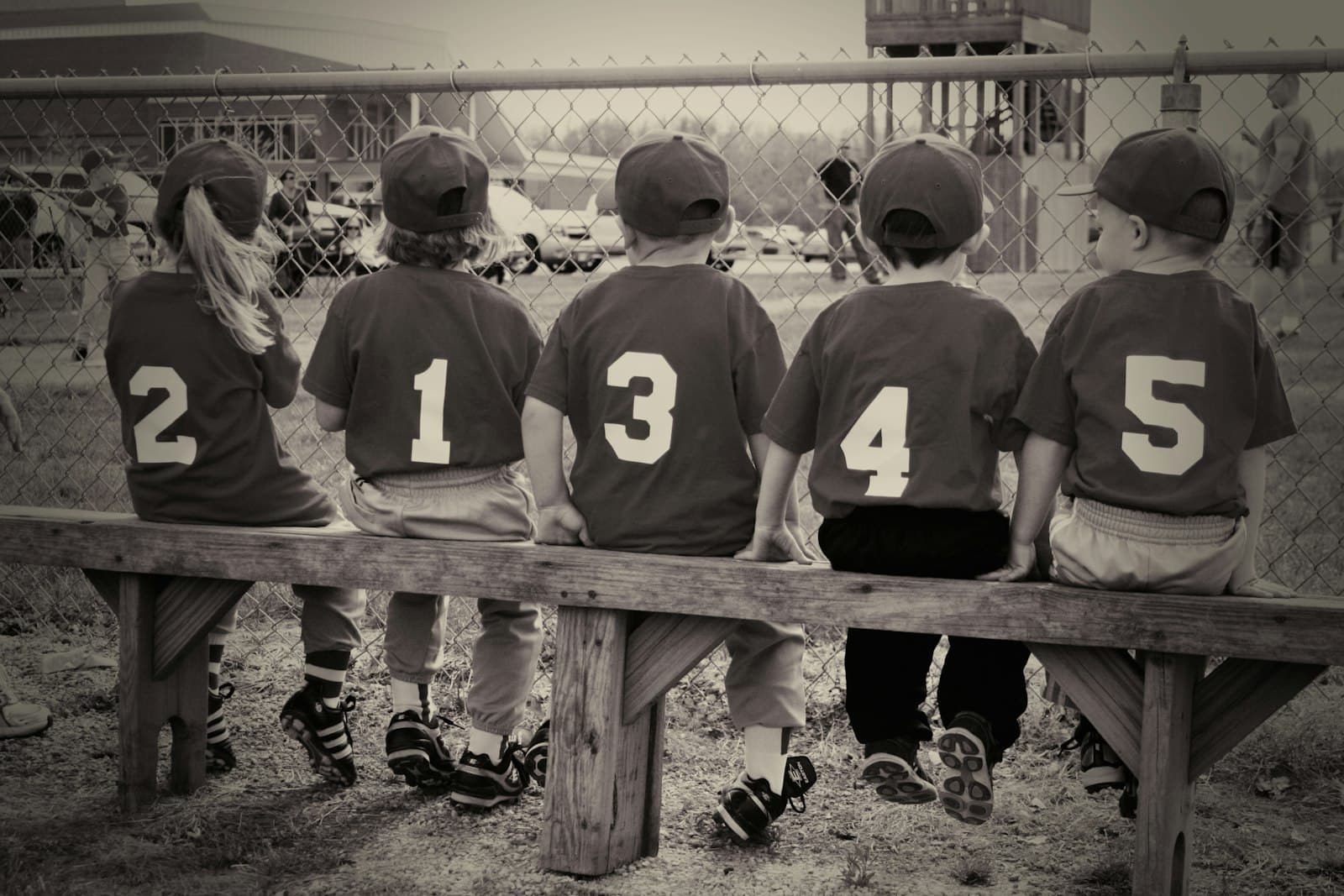 Five kids in numbered jerseys sitting on a dugout bench, seen from behind.